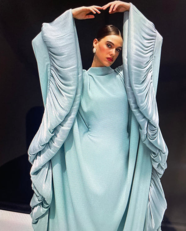 Model wearing a flowing light blue abaya with dramatic pleated sleeves and sides, featuring a high neckline